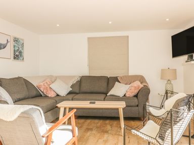 A living room with a grey sectional sofa a wooden coffee table two wire chairs with cushions a wooden armchair a lamp and a wall mounted television at The Beach Halt in Perranporth