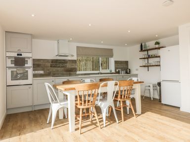 A kitchen with a wooden dining table and mixed chairs a built in oven a hood a sink shelves with glasses and a white fridge at The Beach Halt in Perranporth