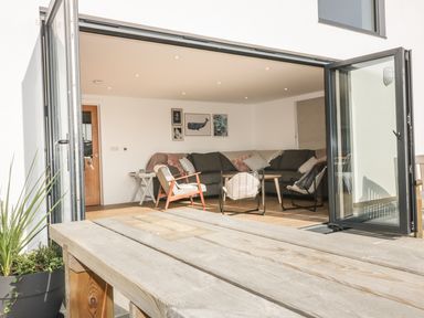 An outdoor wooden table with a large open glass door leading to a living room with dark sofas and framed pictures on the wall at The Beach Halt in Perranporth
