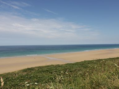 A wide sandy beach with a strip of greenery in the foreground and ocean under a partly cloudy sky at The Beach Halt in Perranporth