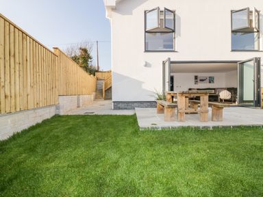 A backyard with green grass wooden fence and patio with wooden table and benches at The Beach Halt in Perranporth