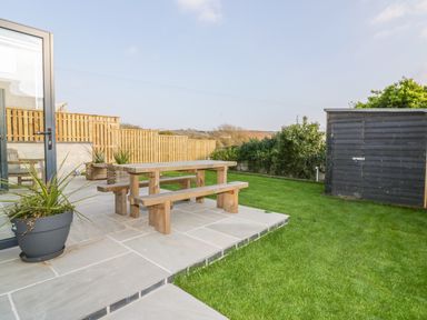 An outdoor patio area with a wooden picnic table and benches a potted plant on tiled flooring a wooden fence surrounding a grassy lawn and a garden shed at The Beach Halt in Perranporth