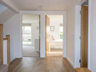 A hallway with wooden floors leading to two rooms with open doors and a staircase with wooden railing at The Beach Halt in Perranporth