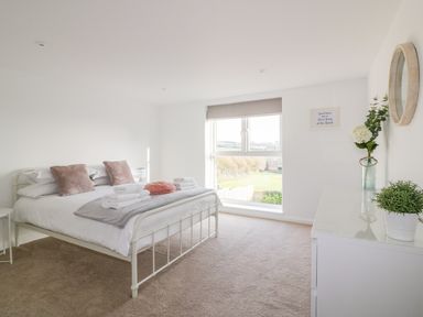 A bedroom with a white metal bed frame, pillows, folded towels, a large window, a white dresser with plants and a round mirror at The Beach Halt in Perranporth