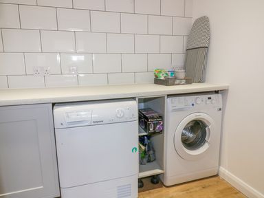 A laundry room with a washing machine and dryer under a countertop with an ironing board and cleaning supplies on top at The Beach Halt in Perranporth