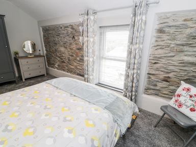 A bedroom with a bed covered in patterned bedding a gray wardrobe a dresser with a round mirror a window with curtains and exposed stone walls at Bryn Alun in Tanygrisiau near Blaenau Ffestiniog