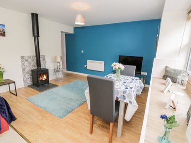 A living room with a blue accent wall a wood stove a dining table with flowers and a blue rug at Bryn Alun in Tanygrisiau near Blaenau Ffestiniog