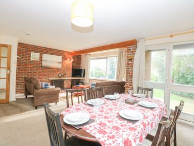 A dining table with red floral tablecloth and set plates and glasses in a living room with brown sofas and brick walls at Swan Dyke Cottage in Horning near Wroxham
