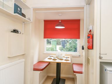 A small dining area with a table and red cushioned benches near a window with a red blind at Swan Dyke Cottage in Horning near Wroxham