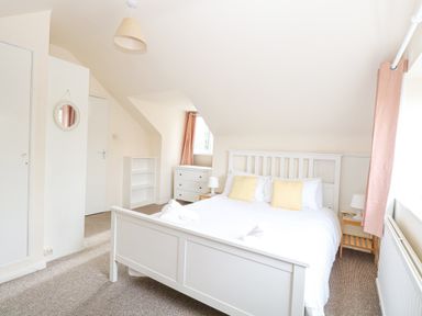 A bedroom with a white bed with yellow pillows a bedside table a chest of drawers a small bookshelf and curtains at Swan Dyke Cottage in Horning near Wroxham