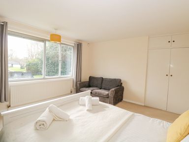 A bedroom with a bed, rolled towels, a gray sofa, window with gray curtains, and built-in white wardrobes at Swan Dyke Cottage in Horning near Wroxham