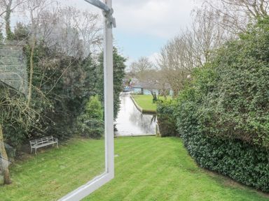 A view of a grass lawn with bushes trees and a river beyond taken through an open window at Swan Dyke Cottage in Horning near Wroxham