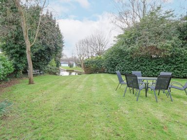 A grassy garden with outdoor table and chairs near trees and a small waterway at Swan Dyke Cottage in Horning near Wroxham