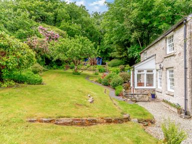A stone cottage with a white porch next to a garden with trees shrubs and a lawn at Westside Cottage in Cardinham near Bodmin