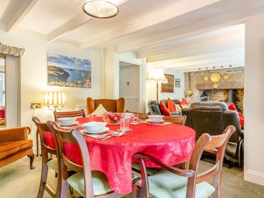 A dining area with a red tablecloth on a round table set for four near a living room with black leather sofas and a stone fireplace at Westside Cottage in Cardinham near Bodmin