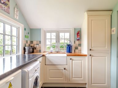 A kitchen with a washing machine a sink with a window above cream cabinets and a fire extinguisher at Westside Cottage in Cardinham near Bodmin