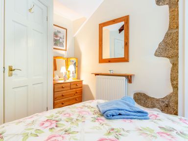 A bedroom with a floral bedspread a wooden dresser with a lit lamp a wall mirror and a folded blue towel at Westside Cottage in Cardinham near Bodmin