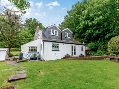 A house with two dormer windows a stone pathway and green lawn surrounded by trees at Westside Cottage in Cardinham near Bodmin