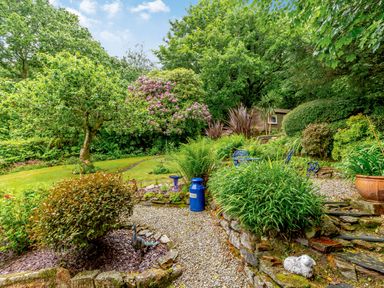 A garden with various plants trees gravel paths blue outdoor furniture and a small wooden shed at Westside Cottage in Cardinham near Bodmin