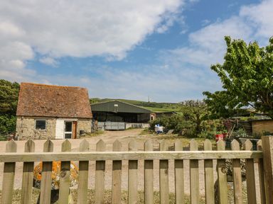 A stone building with a tiled roof behind a wooden fence on a farm with trees and a large shed in the background at Chale Farm Lodge in Chale