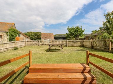 A fenced grassy backyard with wooden steps and a picnic table surrounded by stone and wooden buildings at Chale Farm Lodge in Chale