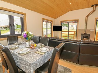 A dining area with a table set for six with dishes and wine next to a living area with a black sectional sofa television and a wood stove at Chale Farm Lodge in Chale