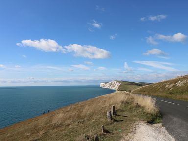 A coastal road with grassy hills and a sea cliff under a blue sky with clouds