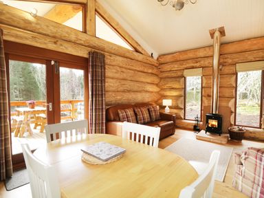 A log cabin living room with a dining table chairs sofa wood stove and glass doors leading to a deck at Heron Lodge in North Kessock