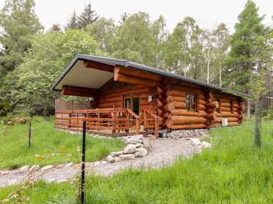 A wooden log cabin with a covered porch and gravel pathway surrounded by grass and trees at Heron Lodge in North Kessock