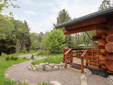 An outdoor view of a wooden cabin porch beside a gravel path and trees at Heron Lodge in North Kessock
