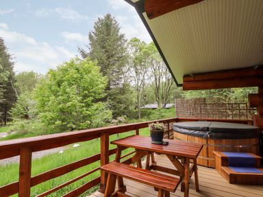 A wooden deck with a picnic table and benches and a covered hot tub surrounded by trees at Heron Lodge in North Kessock