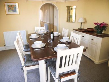 A dining room with a wooden table set for six with white chairs and tableware at Pendeen in Wadebridge