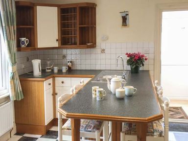A kitchen with a black countertop island with cups and a teapot on it wooden cabinets a sink and a window with curtains at Pendeen in Wadebridge
