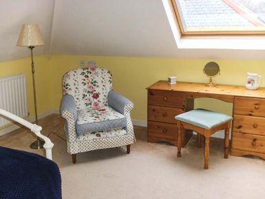 A room with a floral armchair a wooden desk with drawers a small stool a standing lamp and a skylight window at Pendeen in Wadebridge
