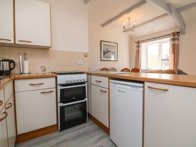 A kitchen with white cabinets a small oven a fridge and a dining area with chairs and a window at The Cottage in Chilsworthy near Holsworthy