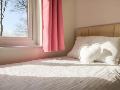 A bed with white linens and a heart-shaped pillow next to a window with pink curtains at The Cottage in Chilsworthy near Holsworthy