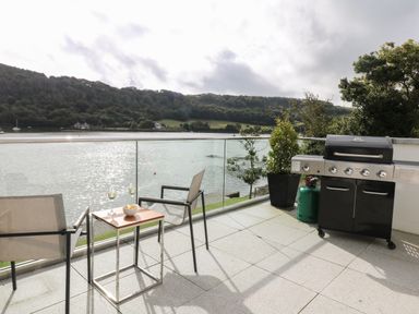 An outdoor terrace with two chairs a small table with snacks and drinks a barbecue and a view of a river and wooded hills at Ancarva Near Kingsands