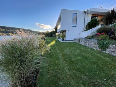 A modern white house with large windows and a grassy lawn next to a body of water at Ancarva Near Kingsands