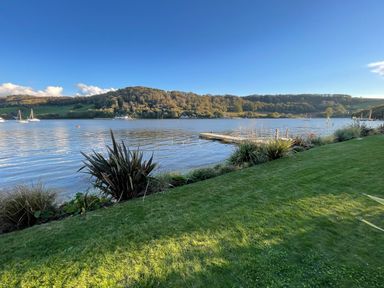 A grassy lawn with plants and a wooden dock by a river with hills and boats in the background at Ancarva Near Kingsands