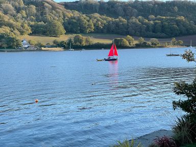 A body of water with boats including one with red sails and a house with trees on the far shore at Ancarva Near Kingsands