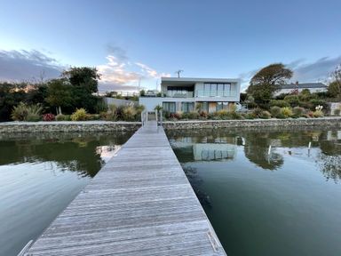 A wooden dock leading to a modern two-story house with glass railings by a body of water at Ancarva Near Kingsands