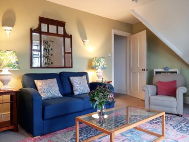 A living room with a blue sofa two patterned cushions a grey armchair with a red cushion a wooden glass coffee table with a vase of flowers and two side tables with lamps at Haslington House in Malvern