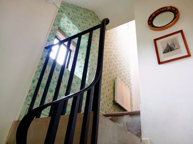 A staircase with black railing wallpapered walls a radiator a window an oval mirror and a framed picture at Haslington House in Malvern