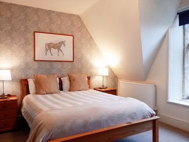 A bedroom with a wooden bed with pillows and a blanket two bedside tables with lamps and a framed horse picture on a patterned wall at Haslington House in Malvern