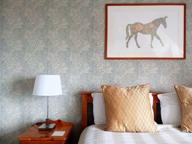 A bedroom with patterned wallpaper a framed horse artwork a wooden bedside table with a lamp and a bed with beige pillows at Haslington House in Malvern