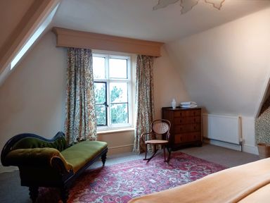 A sitting room with a green upholstered chaise lounge a wooden chair a chest of drawers and a window with floral curtains at Haslington House Malvern