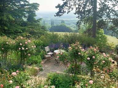 An outdoor garden seating area with wooden chairs and a round table surrounded by flowers and trees at Haslington House in Malvern