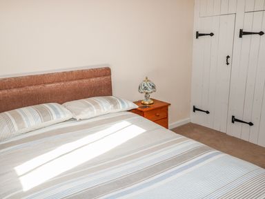 A bedroom with a bed striped bedding a wooden nightstand with a lamp and white closet doors at Honeysuckle Cottage in Ashwater near Holsworthy