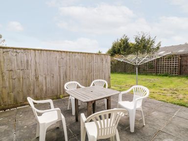 An outdoor patio area with a wooden table surrounded by five white plastic chairs and a rotary clothesline on a paved and grassy yard at Honeysuckle Cottage in Ashwater near Holsworthy