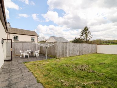 An outdoor patio with a table and four chairs next to a grass lawn and a wooden fence at Honeysuckle Cottage in Ashwater near Holsworthy